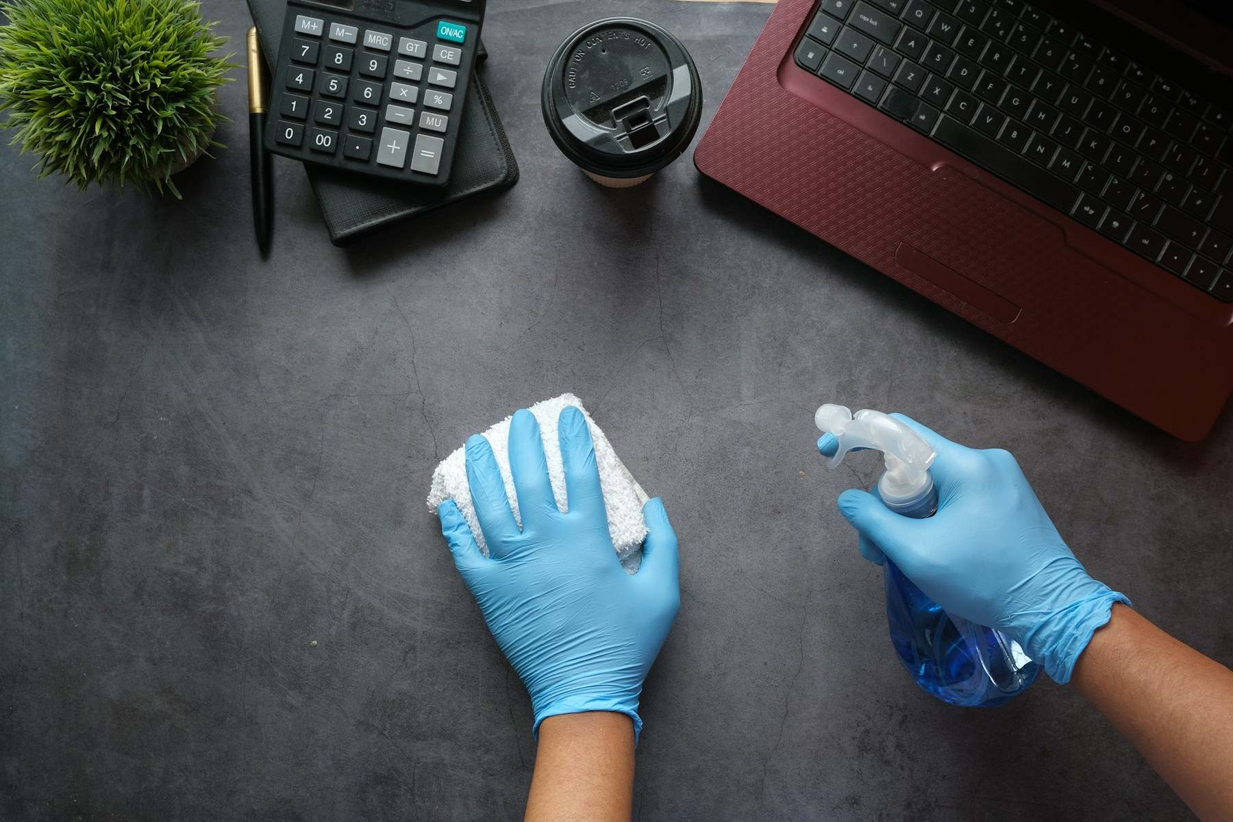 Hände in Handschuhen wischen einen Schreibtisch mit Tuch und Sprühflasche ab