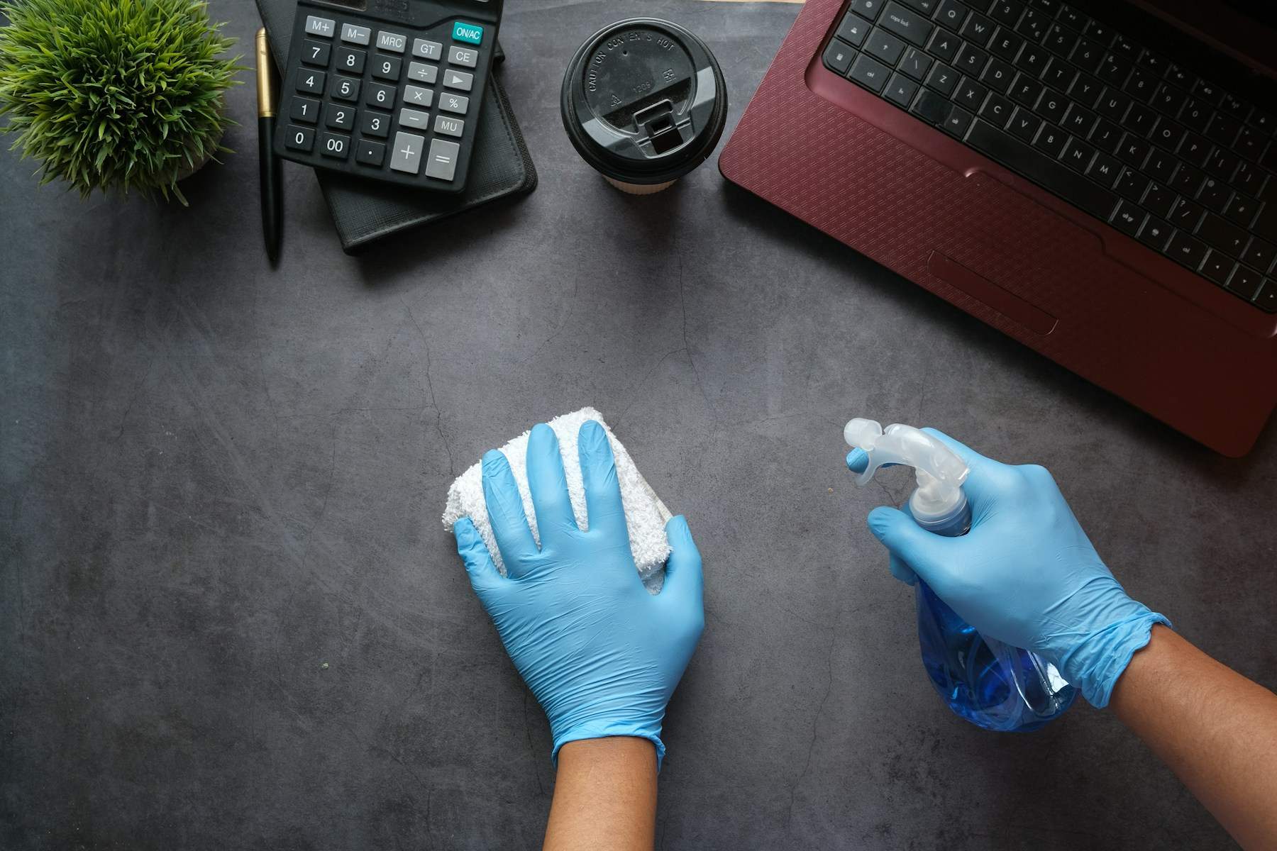 Hände in Handschuhen wischen einen Schreibtisch mit Tuch und Sprühflasche ab